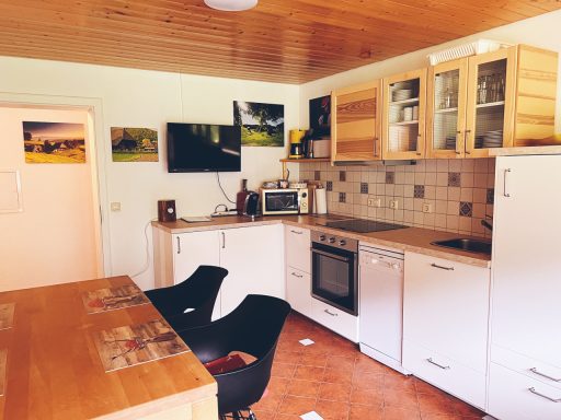 Helle Küche mit Holzregalen, Kühlschrank, Herd und Esstisch. Fotos an der Wand.